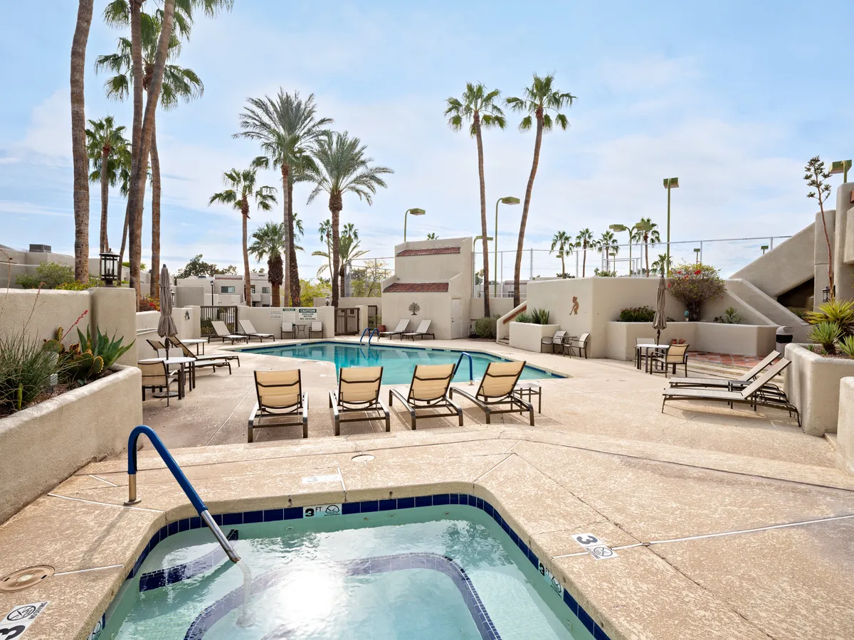 This image displays a sparkling swimming pool surrounded by lounge chairs, a hot tub, and palm trees, capturing the quintessential resort-style relaxation area within a gated community. The inviting pool and spa area is accented by tranquil landscaping and sun-drenched spaces ideal for sunbathing, socializing, and unwinding.