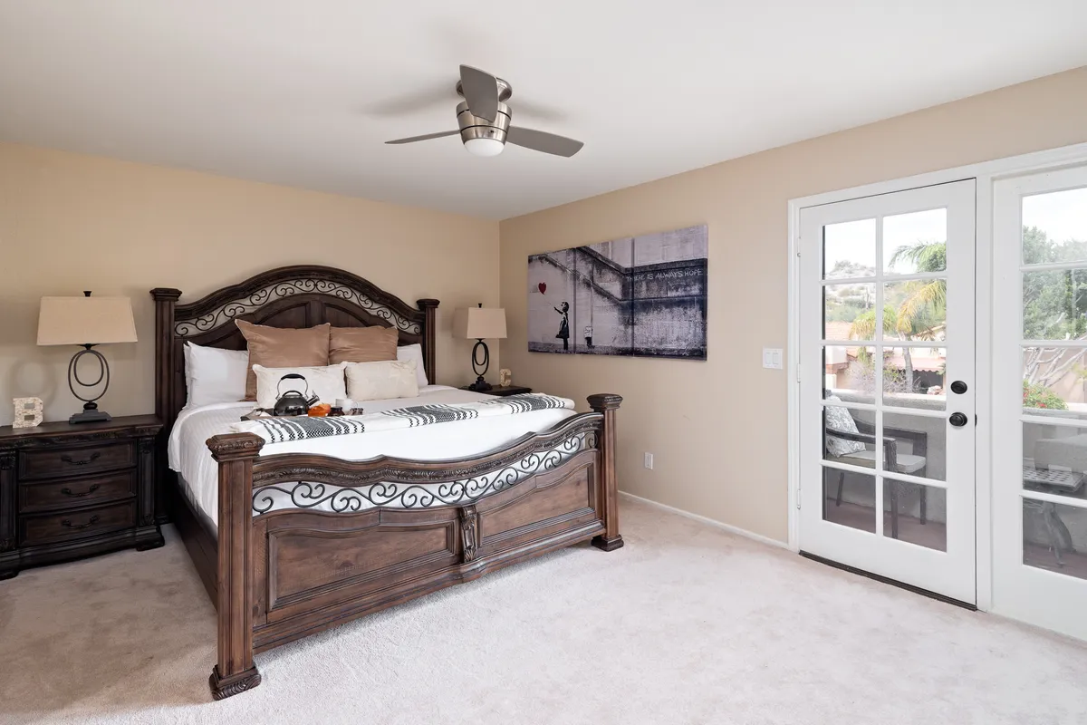 A spacious bedroom featuring a large wooden bed topped with soft linens, inviting pillows, and a serene atmosphere. French doors open to an outdoor balcony, allowing natural light to flood the room. Elegant wall art and subtle décor touches accentuate the restful ambiance, while the overall layout is ideal for relaxation or enjoying a quiet morning coffee inside or outside.