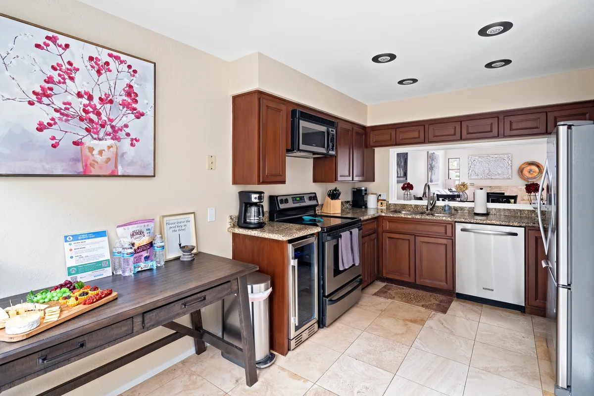 A modern, fully equipped kitchen with sleek appliances, granite countertops, a wooden table set with snacks, and framed artwork on the walls. The open design connects seamlessly with the adjacent living space, offering an ideal setting for meal prep, entertaining, and casual dining.