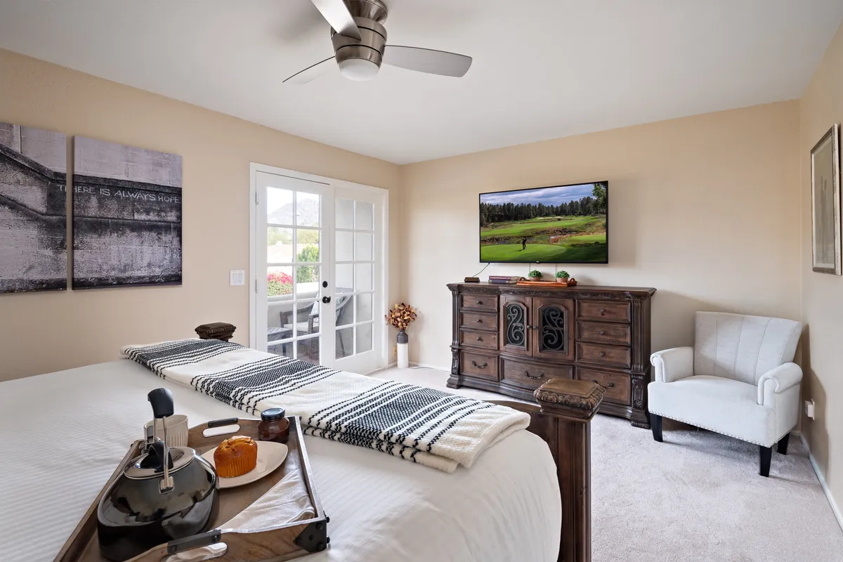 A cozy bedroom with a plush bed adorned with a decorative throw, a wooden dresser, and a wall-mounted TV. French doors lead to an outdoor space, contributing to a sense of openness and connection with nature. The room's soothing color palette and soft textures encourage relaxation, and the presence of the TV adds to the comfort and amenities mentioned for the primary suite.